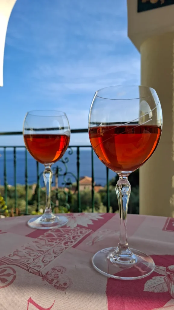 Two wine glasses on a private balcony overlooking the turquoise Aegean Sea and beach, an invitation to stay at our Agia Paraskevi Studios.