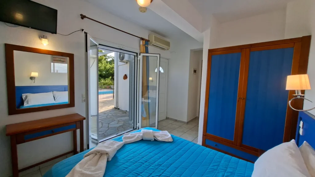 Bright bedroom looking out through open doors to a sunlit terrace and swimming pool at these studios with pool Greece.