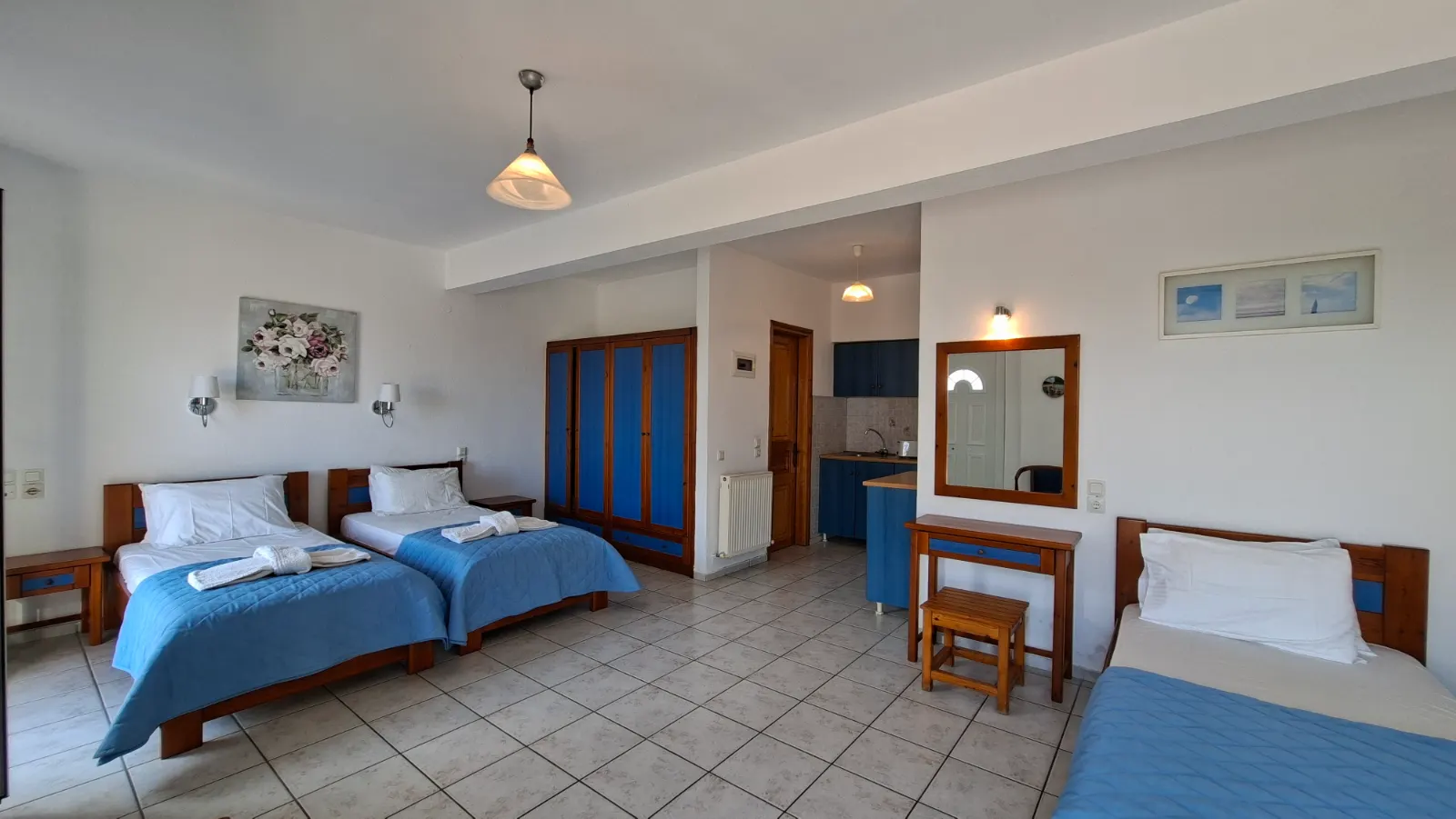 Full, slightly angled interior view of the Triple Studio showing all three single beds, the large blue wardrobe, and the recessed kitchenette area.